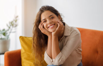 A woman with long brown hair, wearing a beige shirt, sits on an orange sofa and smiles at the camera with her hands resting on her face.