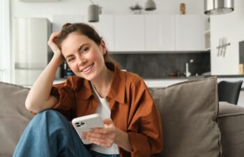 Portrait of cute young smiling woman, sitting on sofa with mobile phone, relaxing at home and using smartphone, scrolling social media, buying online, ordering something on application.